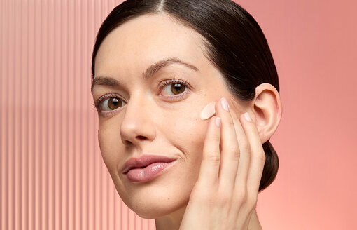 woman applying cream to her cheek on a peach background